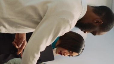 Dikey video: Asyalı çalışanın müşteriye giysi mağazasında denemesi için gömlek vermesi, müşteriye modern kıyafetler alması için yardım etmesi. Genç bir adamla yeni moda koleksiyonu ve küçük işletmeler hakkında konuşan bir kadın.