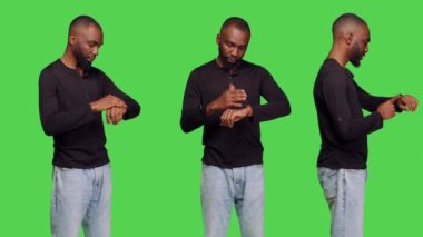 Kol saatini kontrol eden genç, dakik bir yetişkin, yeşil ekranın arkasında duruyor. Kot pantolon giyen ve saati olan bir erkek, kameraya geç kalıyor. Rahat bir poz..