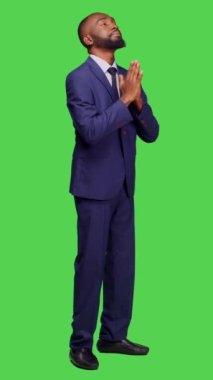 Dikey video: Girişim girişimcisinin stüdyoda iyi şanslar dilemek için yeşil perde arkaplanının üzerine resmi takım elbise giymesinin yan görüntüsü. Genç bir erkek işçi dua işareti ile el ele tutuşuyor, dindar.