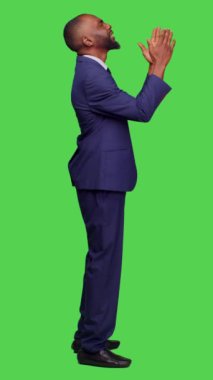 Dikey video: Girişimci profili stüdyoda iyi şanslar dilemek için Tanrıya dua ediyor, yeşil perde arkaplanı üzerine resmi takım elbise giyiyor. Genç bir erkek işçi dua işareti ile el ele tutuşuyor, dindar.