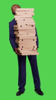 Dikey video: Erkek girişimcinin kameraya büyük pizza kutuları yığını tutarken ön görüntüsü, tüm vücudun üzerinde fast food siparişi taşıyor. Takım elbiseli genç adam yemek hazırlıyor.