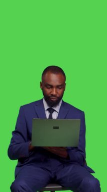 Dikey video: Stüdyoda dizüstü bilgisayarda çalışan, kablosuz bilgisayarla koltukta oturan Afro-Amerikan yöneticinin ön görüntüsü. Ofis çalışanının takım elbisesi yeşil yerine taşınabilir bilgisayar kullanıyor.