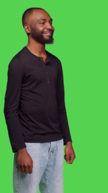 Dikey video: Stüdyoda yeşil ekran arkaplanında poz veren, kamerada arkadaş canlısı ve iyimser bir görünüm. Kot pantolonlu ve gömlekli genç bir yetişkin stüdyoda pozitif çıkıyor.