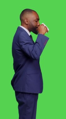 Dikey video: Stüdyoda kahve fincanı içen genç bir şirket yöneticisinin profili, yeşil ekran arkaplanı üzerinde duruyor. İş adamı kafein içeceğini ya da içeceğini içiyor, iyimser.