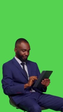 Dikey video: Stüdyoda sandalyede oturan dijital tablet kullanarak sosyal medya sayfasında kayan erkek girişimcinin yan görüntüsü. Modern aygıtlarda online ağ taraması yapan genç girişimci