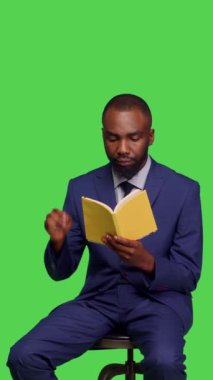 Dikey video: Stüdyoda edebiyat kitabı okuyan modern girişimcinin koltukta oturan takım elbise giymesinin ön görüntüsü. Olumlu iyimser adam yeşil ekranda ders veriyor.
