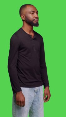 Dikey video: Genç erkek modelin görüntüsü kamerada şık poz veriyor, yeşil ekran arkaplanının üzerinde duruyor. Kot pantolon ve gömlek giyen, kendine güveni olan, pozitif yüz ifadesi olan sıradan biri.