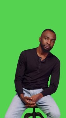 Dikey video: Afro-Amerikan bir adamın sandalyede oturup beklemesi, kendine güvenmesi ve şık kot ve tişörtü ile rahat hissetmesi. Erkek model stüdyoda iyimser ve arkadaş canlısı davranıyor.