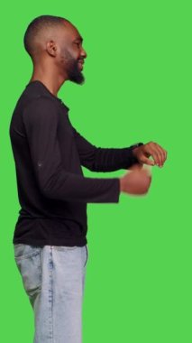 Dikey video: Kol saatinde, yeşil ekran arkaplanının üzerinde duran dakik genç yetişkin kontrol zamanı profili. Kot pantolon giyen ve saati olan bir erkek, kameraya geç kalıyor. Gelişigüzel