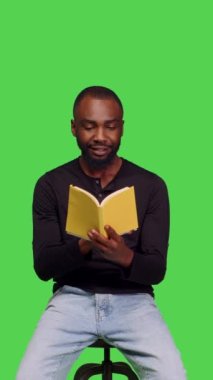 Dikey video: Yeşil ekran stüdyosundaki sandalyede oturan ve kamerada sayfalar okumak için kendine güvenen yetişkinlerin kitap tutuş görüntüsü. Erkek model edebiyat öyküsü ya da boş zaman aktivitesi için roman