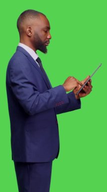 Dikey video: Stüdyoda çalışması için dijital cihaz tutan ofis yöneticisinin profili, sosyal medya uygulaması üzerinden Greescreen arka planında geziniyor. Modern tablet kullanan resmi takım elbiseli bir şirket çalışanı