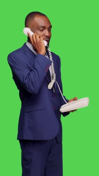 垂直视频 美国黑人男子用固定电话给经理打电话的侧视图 用电线接听办公室电话 在工作室里进行远程聊天 男性员工在绿屏背景下摆姿势 — 图库视频影像