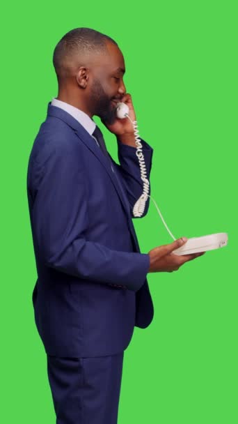 垂直ビデオ スタジオでのリモートチャットのためのコードでオフィスの電話に応答し マネージャーを呼び出すために固定電話を使用してアフリカ系アメリカ人の男のプロフィール 緑の画面の背景にポーズをとる男性従業員 — ストック動画