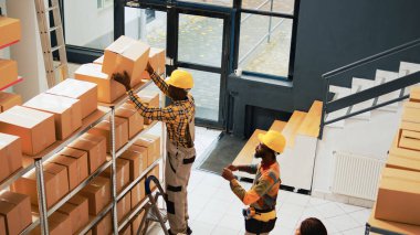 Group of workers working on stock inventory in depot, organizing packages and boxes on racks and shelves. Team of people moving cardboard containers in storage room. Handheld shot.