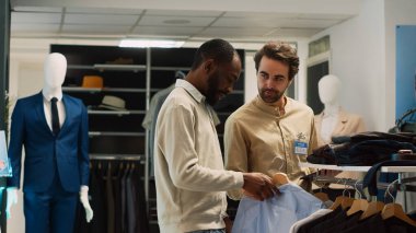 Genç alışverişçi, mağaza çalışanından kıyafet için yardım istiyor. Moda mağazasından resmi giysiler almak istiyor. Perakende satış görevlisi müşteriyle modern kıyafet alışverişi hakkında konuşuyor..