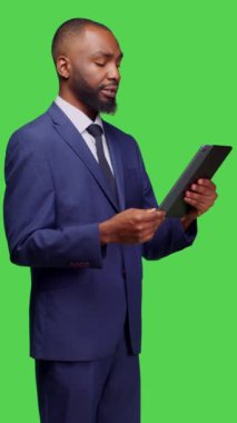 Dikey video: Kamerada çalışmak için dijital tablet tutan erkek çalışanın yan görüntüsü, sosyal medyada yeşil ekran arkaplanı üzerinden kayıyor. Yeni başlayanlar için ofis kıyafeti, modern aygıt, gezinme