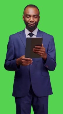 Dikey video: Kamerada çalışmak için dijital tablet tutan erkek çalışanın ön görüntüsü, yeşil ekran arkaplanı üzerinden sosyal medyada kayıyor. Modern cihazlar kullanarak ofis elbisesi giyen bir girişimci