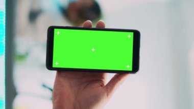 Dikey video: Genç çalışan yeşil ekranlı cep telefonu kullanıyor, akıllı telefondan izole edilmiş telif alanı görüntülerini gösteriyor. Erkek mağaza asistanı butikte boş krom anahtar şablonu tutuyor. Kapat