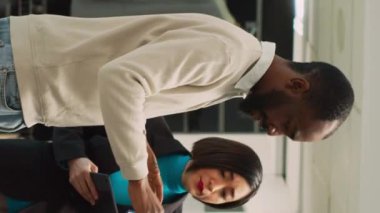 Dikey video: Alışveriş yapmak için raflarda giysileri inceleyen çeşitli insanlar, alışveriş merkezinde mağaza asistanı olarak çalışan bir kadın erkeğin gömlek seçmesine yardım ediyor. Moda ürünlerine bakan bir müşteri. El bilgisayarı