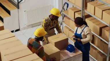 Afro-Amerikalı çalışanlar envanter ve lojistik üzerinde çalışıyorlar. Ürünleri sevkiyata hazırlamak için panodaki hisse listesine bakıyorlar. Depo çalışanları ürünleri kutularda kontrol ediyor.