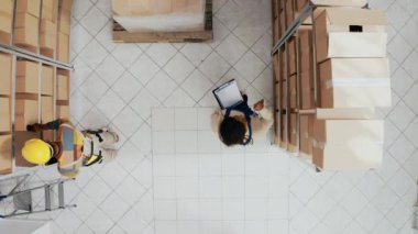 Depoda kalite kontrol yapan bir grup insan karton paketlerle dolu raflar ve raflar arasında duruyor. Kutular ve kargolarla çalışan çalışanlar. İHA atışı.