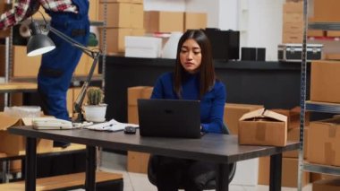 Asian owner trying to improve budgeting on laptop in depot, checking merchandise stock and inventory on pc. Female entrepreneur working on business development with distribution orders.