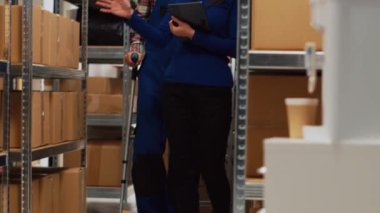 Koltuk değnekleriyle yürüyen, işyeri sahibiyle konuşan, depo alanında ürün envanteri yapmak için tablet kullanan bir adam. Çalışan bir kadınla mal hakkında konuşuyor, fiziksel bir sorun. El kamerasıyla..