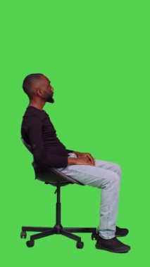 Dikey video: Afro-Amerikan kişinin profili sandalyede bir şey bekliyor, tüm vücudu yeşil ekranda oturuyor. Genç erkek model kamerada kendinden emin ve iyimser hissediyor.