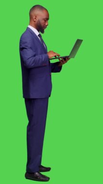 Dikey video: Modern çalışan profili ağı taramak için dizüstü bilgisayar kullanıyor, çevrim içi siteye tam vücut yeşil ekranının başında duruyor. Genç iş adamı kablosuz bilgisayarla çalışıyor.