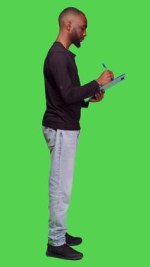Dikey video: Erkek model profili pano kağıtlarına not alıyor, tüm vücudun yeşil ekranının başında duruyor. Genç, neşeli yetişkin, kamerada pozitifken boş zamanlarında dosyalara bilgi yazıyor.