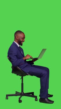 Dikey video: İş planı üzerinde çalışmak için dizüstü bilgisayar kullanan şirket çalışanlarının profili, stüdyoda resmi takım elbise giyen sandalyeye oturmaları. Genç yönetici kablosuz bilgisayarda internet sayfasına göz atıyor, tam vücut