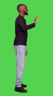 Dikey video: Afrikalı Amerikalı bir adamın stüdyodaki insanlara el sallayarak selam verişi ya da kamerada veda edişi. Genç model gülümsüyor ve yeşil ekran arkasında duran birini selamlıyor.