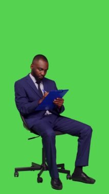 Dikey video: Afro-Amerikan iş adamının dosyalara not alıp yeşil perde arkaplanı üzerine yeni kurumsal fikirleri düşünmesi. Erkek girişimci kağıtlara iş bilgileri yazıyor