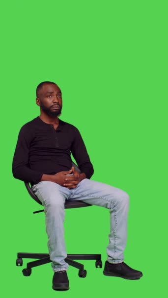 垂直视频 非洲裔美国人在椅子上等东西的侧视图 坐在全身绿色的屏幕背景上 年轻男模在镜头前感到自信和乐观 — 图库视频影像