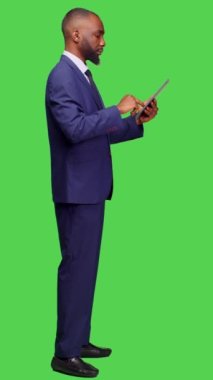 Dikey video: Tablet üzerinde internet sitesine göz atan modern çalışanın profili, tam vücut yeşil ekran arkaplanı üzerinde çevrimiçi ağ sayfası ile çalışıyor. Ofis takımında genç bir yetişkin, elinde aletle.