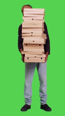 Dikey video: Kameraya büyük bir pizza yığını teslim eden, içinde fast food siparişi olan karton kutular taşıyan bir gencin ön görüntüsü. Tüm vücudu yeşil ekranda poz veren paketlerle yetişkin bir erkek.