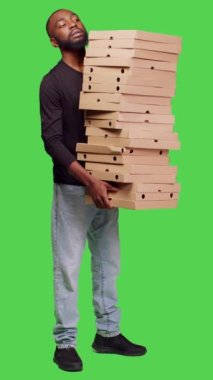 Dikey video: Kameraya büyük bir pizza yığını teslim eden, fast food siparişleriyle dolu karton kutular taşıyan genç insanların yan görüntüsü. Tüm vücudu yeşil ekranda poz veren paketlerle yetişkin bir erkek.