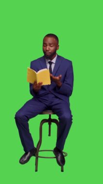 Dikey video: Pozitif çalışanın stüdyoda oturup, yeşil ekran arka planında edebiyat okurken mutlu hissettiği masal kitabının ön görüntüsü. Sandalyedeki genç iş adamı