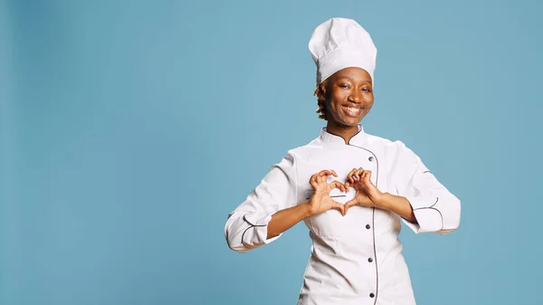Female cook showing heart shaped symbol on camera, expressing romantic gesture and wearing cooking apron. Young adult chef being flirty with romance love sign, culinary industry.