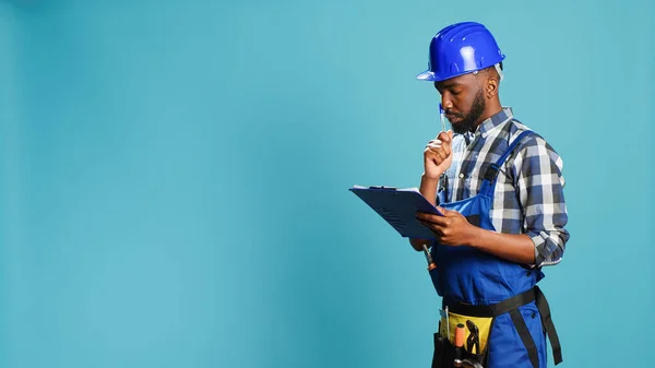 Confident builder making calculations on papers, taking notes on files before starting professional construction project in studio. Male carpenter writing information and measurements.