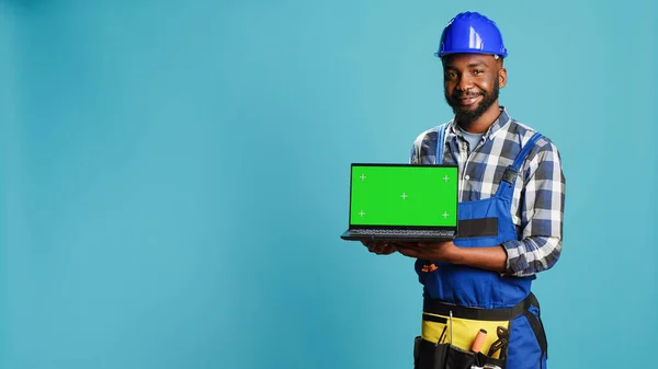 Smiling craftsman showing laptop with green screen on camera, advertising modern isolated display on pc. Young builder contractor holding computer with blank chroma key copyspace screen.