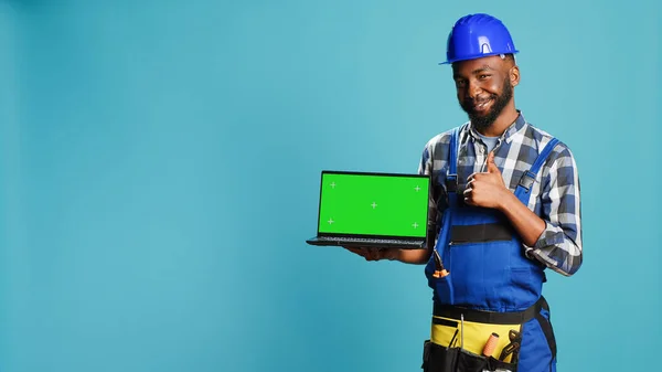 Happy mechanic pointing at laptop with greenscreen on camera, advertising isolated template display on pc. Young construction worker showing computer with chroma key mockup copyspace.