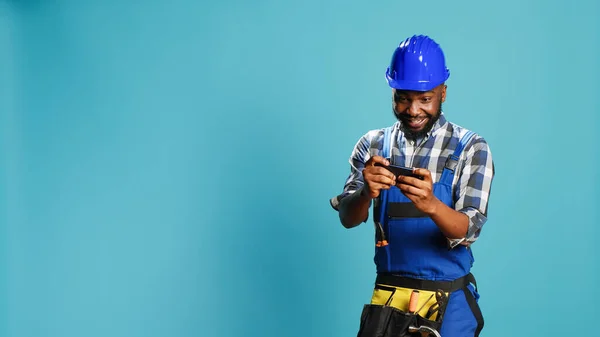 Confident repairman playing mobile video games over blue background, having fun with online gaming competition in studio. Young professional builder enjoying game play challenge.