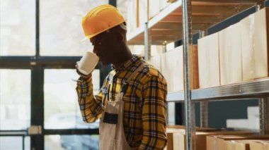 Male storage room employee standing near racks filled with goods in cardboard boxes. Young worker with overalls working in warehouse with retail store merchandise, transportation order.