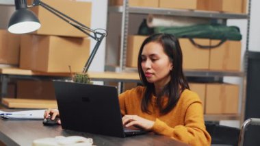 Female owner working in wheelchair at warehouse, using laptop to plan logistics and distribution in depot. Woman with chronic disability doing financial planning and quality control. Handheld shot.