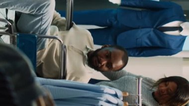 Dikey video: Tekerlekli sandalyedeki genç müşteri kıyafet alışverişi yapıyor, moda mağazasından günlük giysiler alıyor. Perakende satış malına bakan kronik arızalı bir adam var.