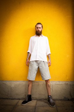 Sakallı hippi, sarı duvardaki kameraya bakıyor.