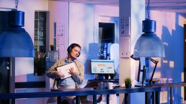 Happy advisor working on business report with headset, listening to music and checking economy diagrams on pc. Female worker planning startup progress before deadline, wearing headphones.