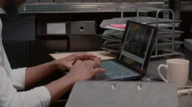 Afro-Amerikalı bir adam laptopta fotoğrafları inceliyor, olay mahalli gözetleme materyallerini olay odasına koyuyor. Profesyonel hukukçu, duvardaki kanıt panosunu inceliyor..