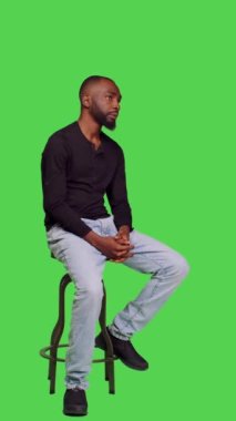 Dikey video: Sandalyedeki Afro-Amerikan adam özgüvenle poz veriyor, tüm vücudu yeşil ekranda oturuyor. Genç erkek model kendini iyimser hissediyor ve stüdyoda bir şeyler bekliyor.